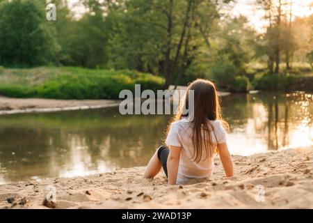 Adorable petite fille aux cheveux sombres et longs confortables assis sur le sable près de la forêt. Enfant féminin portant un t-shirt blanc et un short noir jouant avec Banque D'Images