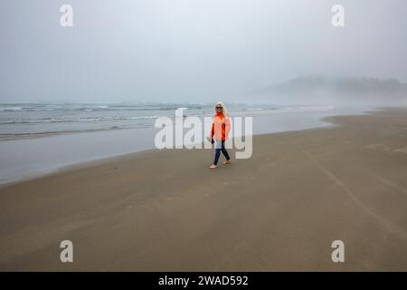 Femme en veste orange marche le long de la plage brumeuse Banque D'Images