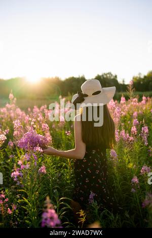 Vue arrière de la femme dans le chapeau de paille debout dans la prairie au coucher du soleil Banque D'Images