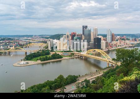 La ligne d'horizon du centre-ville de Pittsburgh, un pont en arc jaune et une large rivière bordée d'arbres verts se trouvent sous un ciel nuageux stratifié en Pennsylvanie. Banque D'Images