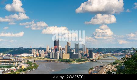 La ligne d'horizon de Pittsburgh s'étend sur une rivière parsemée de bateaux sous de gros nuages cumulus blancs, avec des feuilles en bas à droite, Pennsylvanie. Banque D'Images