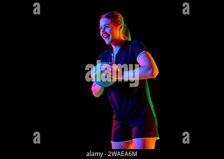 Portrait de jeune femme, joueur de handball tenant la balle pour faire parfait servir précis tôt dans le jeu sur fond noir dans la lumière néon. Banque D'Images