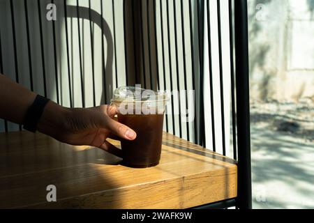 Main tenant le café de glace noire dans le verre à boire sur fond de table en bois avec un rayon de soleil de la fenêtre. Boisson rafraîchissante d'été. Café noir dans le Banque D'Images