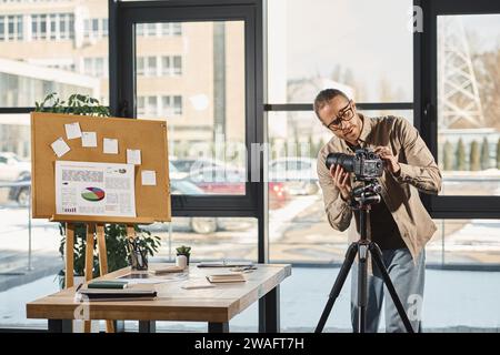 homme d'affaires dans les lunettes ajustant l'appareil photo numérique près du bureau et du liège avec des graphiques dans le bureau Banque D'Images