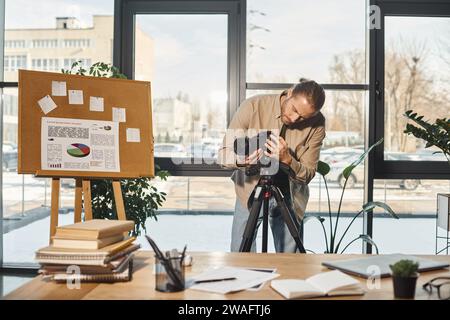 homme d'affaires créatif ajustant l'appareil photo numérique près du tableau de conférence avec des analyses et un bureau de travail dans le bureau Banque D'Images