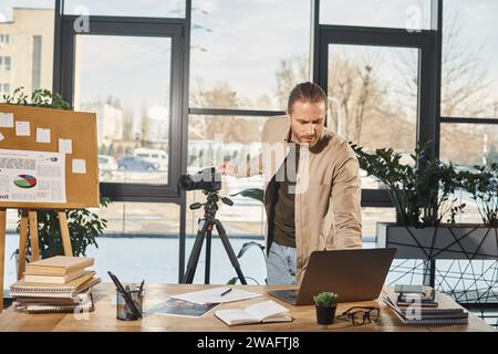 gestionnaire en tenue décontractée ajustant ordinateur portable et appareil photo numérique près du bureau de travail dans le bureau, blogueur vidéo Banque D'Images