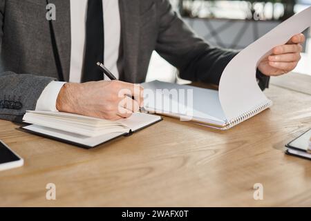 vue recadrée de l'homme d'affaires écrivant dans un cahier sur un bureau de travail dans un bureau moderne, style de vie occupé Banque D'Images