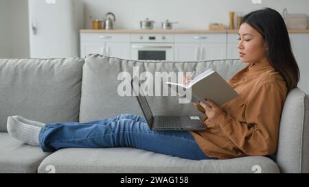 Vue de côté fille asiatique femme sur le canapé avec l'étudiant d'ordinateur portable écrivant des notes dans le copybook se préparant à l'examen faire des devoirs de travail de la maison regarder Banque D'Images