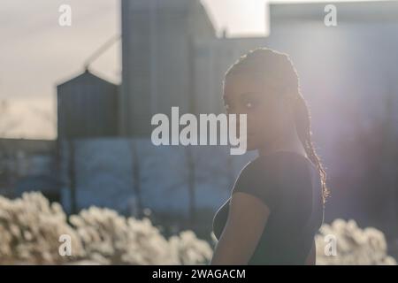 Cette image capture une jeune femme noire jetant un regard sur son épaule, son visage doucement illuminé par la douce lueur d'un soleil couchant. Derrière elle, les silhouettes des bâtiments de la ville s’élèvent contre le ciel, créant une toile d’aspiration urbaine. Le rétroéclairage chaud crée un halo autour de sa silhouette, donnant à la scène une qualité onirique et évoquant un sentiment de contemplation optimiste vers l'avenir. City Glow : Sunset ambiance et Urban Dreams. Photo de haute qualité Banque D'Images