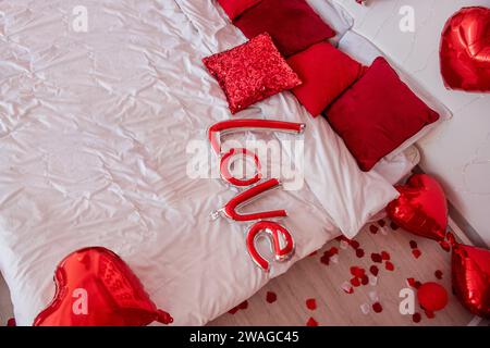 Vue de dessus du ballon en forme d'inscription Love se trouve sur le lit blanc parmi les oreillers rouges. Surprise pour la Saint Valentin. Pétales de rose sur le sol. Placer pour le texte Banque D'Images