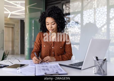 Femme d'affaires expérimentée mature remplissant des formulaires et des tables tout en étant assise au lieu de travail à l'intérieur du bureau, comptable financier sérieux et concentré au travail, enregistrant des données dans des rapports. Banque D'Images