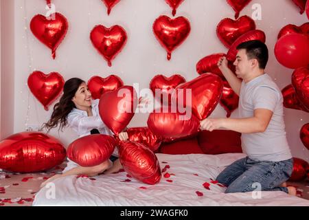 Moment ludique entre un couple engagé dans des ballons d'oreiller combat sur le lit, avec des ballons rouges en forme de cœur en arrière-plan. La femme se trompe joyeusement Banque D'Images