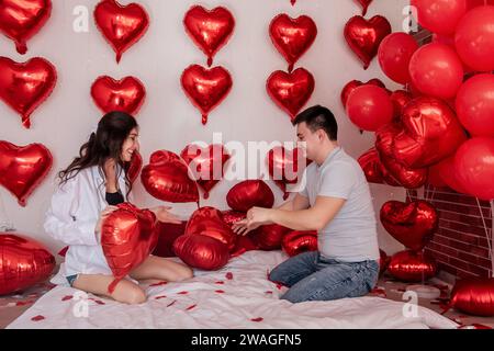 Moment ludique entre un couple engagé dans des ballons d'oreiller combat sur le lit, avec des ballons rouges en forme de cœur en arrière-plan. La femme se trompe joyeusement Banque D'Images