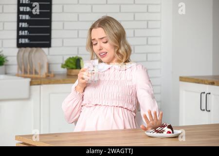 Jeune femme enceinte avec de l'eau et un morceau de gâteau souffrant de toxicose à table dans la cuisine Banque D'Images