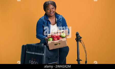 Courrier afro-américain féminin transportant doucement le panier de fruits du sac de livraison de nourriture. Femme de livraison excitée sur son vélo saisissant une boîte de produits frais et sains pour la livraison. Banque D'Images