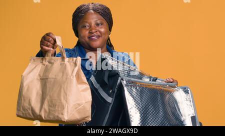 Tout en livrant des repas à emporter sur le vélo, jeune femme noire donnant les pouces vers le haut pour assurer la satisfaction du client. Dans le quartier local, le livreur afro-américain propose une option de dîner sur place écologique. Banque D'Images