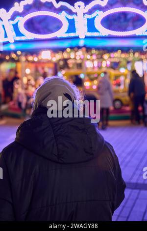 Arrière d'une vieille femme aux cheveux gris avec un foulard devant un carrousel pour enfants illuminé de couleurs Banque D'Images