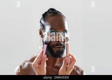 African gay appliquant du maquillage tenant la brosse de maquillage dans la main. Gros plan portrait homme homosexuel regardant l'appareil photo prêt pour appliquer le visage sur le visage. Concept lgbt de la mode. Banque D'Images