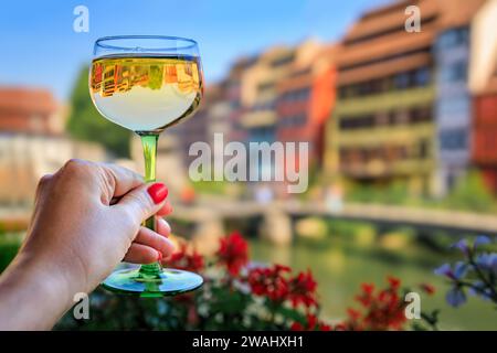 Main de femme avec un verre de vin Gewurztraminer dans un restaurant en plein air, maisons à colombages floues, quartier de la petite France à Strasbourg, France Banque D'Images