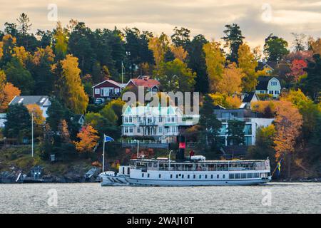 Stockholm, Suède. Banlieues et maisons résidentielles sur les îles à l'est de la ville aux couleurs d'automne. Un bateau de banlieue passant par là. Arbres colorés. Banque D'Images