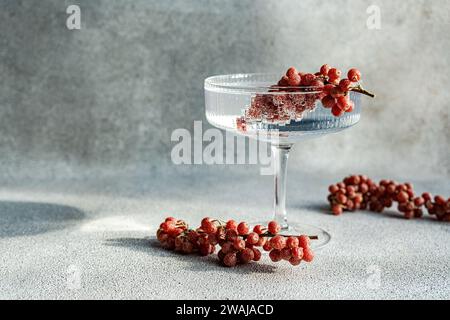 Un élégant verre à cocktail rempli de vin perché sur un fond gris texturé, entouré de grappes éparpillées de raisins rouges Banque D'Images