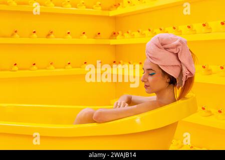 Femme avec une serviette de tête allongée dans une baignoire jaune vibrante entourée d'un motif de canards en caoutchouc miniatures Banque D'Images