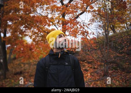 Jeune homme barbu sérieux dans des vêtements chauds avec bonnet de beanie jaune et sac à dos debout près des arbres d'automne colorés dans le parc national de la ville de Stockholm dans da Banque D'Images