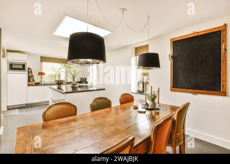 Lampes suspendues suspendues au-dessus d'une table en bois et de chaises avec tableau noir vide dans la salle à manger avec cuisine moderne à la maison Banque D'Images