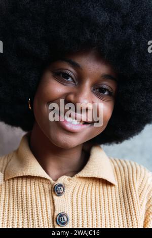 Portrait de jeune afro-américaine gaie aux cheveux maurous et sombres souriant avec joie tout en regardant l'appareil photo contre un mur gris flou Banque D'Images