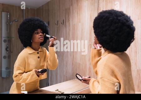 Vue latérale de la jeune femme afro-américaine avec une poudre d'application de coiffure afro avec brosse tout en se tenant devant le miroir dans la salle de bains à la maison Banque D'Images