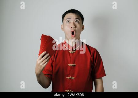 Visage choqué de l'homme asiatique portant Cheongsam ou tissu traditionnel chinois tout en attisant son auto en utilisant angpao ou cadeau monétaire rouge sur fond blanc Banque D'Images