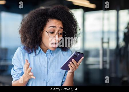 Choqué jeune femme afro-américaine assise dans le bureau et regardant bouleversé à l'écran du téléphone. Gros plan. Banque D'Images