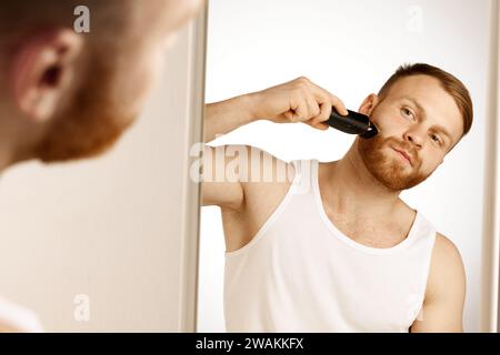 Homme en T-shirt dans la salle de bain se tient devant le miroir pour le toilettage du visage, le bien-être et les soins de la peau à la maison. Banque D'Images