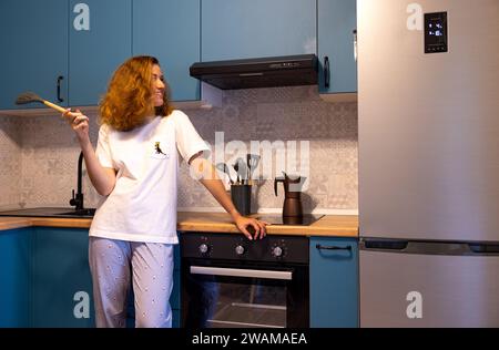 Une jeune fille se tient debout dans la cuisine en pyjama, souriant et tenant une louche Banque D'Images