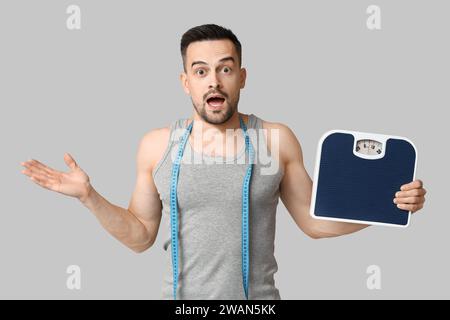 Bel homme sportif surpris avec ruban à mesurer et échelles sur fond gris. Concept de perte de poids Banque D'Images