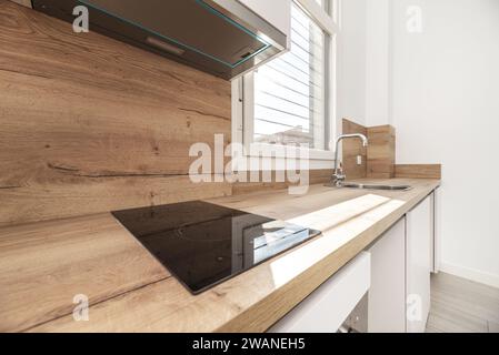 Une cuisine ouverte avec armoires blanches et comptoirs en bois avec dosseret assorti avec petite table de cuisson noire et fenêtre en aluminium blanc Banque D'Images