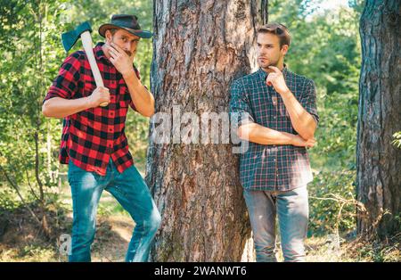 Coupe-bois bûcherons.Vêtements taille basse pour hommes sur le visage sérieux avec hache.Bûcheron brutal et barbu tient hache.Deux bûcherons en forêt.Homme dans le chapeau regarde Banque D'Images