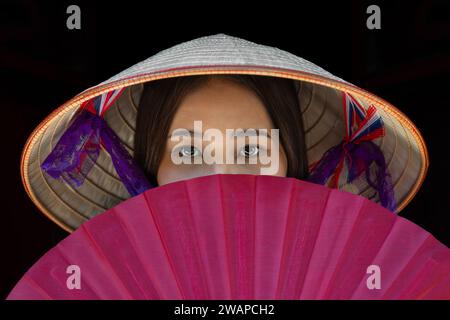 Portrait d'une vietnamienne portant un chapeau conique avec un éventail rouge couvrant sa bouche, Hanoi, Vietnam Banque D'Images