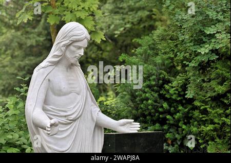 Statue blanche du Christ avec les mains tendues devant le feuillage vert Banque D'Images