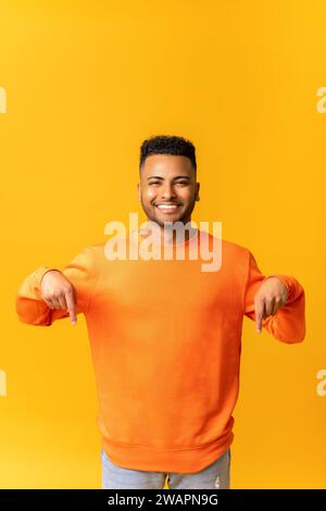 Homme indien attrayant avec une expression faciale joyeuse pointant vers le bas avec les deux doigts, présentant l'espace de copie. Studio intérieur tourné isolé sur fond orange Banque D'Images