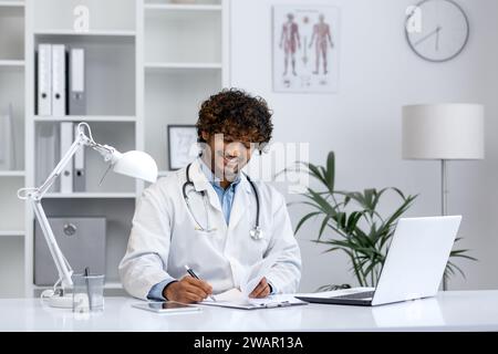 Jeune médecin travaille avec un ordinateur portable à l'intérieur du cabinet médical de la clinique, l'homme sourit joyeusement et remplit des rapports médicaux et des formulaires de traitement, travaille avec des papiers. Banque D'Images