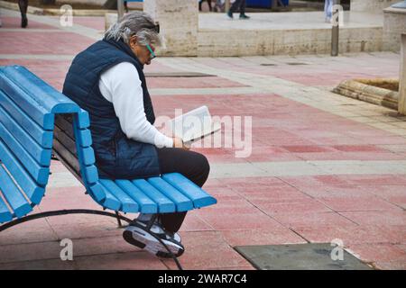 Un blanc caucasien d'âge moyen assis sur un banc de parc public lisant un livre Banque D'Images