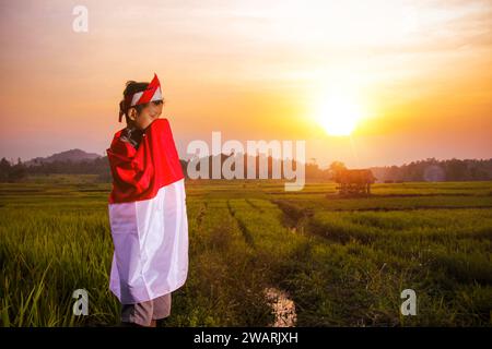 Adolescent asiatique dans les vêtements traditionnels javanais debout tenant le drapeau indonésien et embrassant avec sagesse sur fond de champ de riz vert Banque D'Images