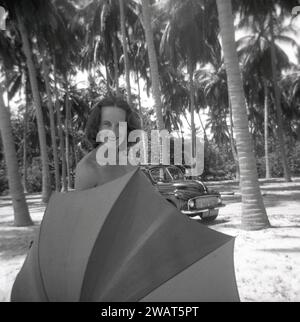 1953, historique, une jeune femme en vacances sur l'île de Ceylan (Sri Lanka) tenant un parapluie pour protéger sa modestie, une voiture de l'époque en arrière-plan garée près des palmiers. Banque D'Images