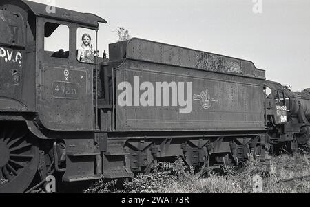 1975, historique, une jeune fille debout dans la cabine du conducteur d'une vieille locomotive à vapeur, non, 4920 à la casse Woodham Brothers à Barry Docks, pays de Galles du Sud, communément connu sous le nom de casse Barry. Derrière le wagon à charbon de la locomotive, locomotive à vapeur 92240, avec le marquage vendu écrit sur le côté. Les Woodham Brothers, négociants en ferraille établis en 1892, apportèrent les anciennes locomotives à vapeur Briitsh Railways, puis les vendirent ou les ferraillèrent. On estime que plus de 200 locomotives à vapeur ont été secourues pour le mouvement de préservation du chemin de fer. Banque D'Images