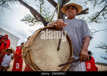 Un homme âgé joue du tambour pendant la danse traditionnelle Yonna. Un groupe Wayuu de filles et un garçon dansent la Yonna pour les touristes qui visitent leur maison à Playa Camarones, à la Guajira, une zone désertique dans le nord de la Colombie. La Yonna, danse traditionnelle du peuple indigène Wayuu, est exécutée pour des célébrations, pour le Majayut (la transition d'une fille indigène à une femme), ou à des occasions liées à la spiritualité. Les femmes portent des robes traditionnelles rouges, tandis que les hommes portent une tenue typique appelée 'wayuuco'. Banque D'Images