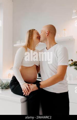 Couple semble très heureux. Un beau mari embrasse sa belle femme enceinte à côté de la fenêtre du salon. Banque D'Images