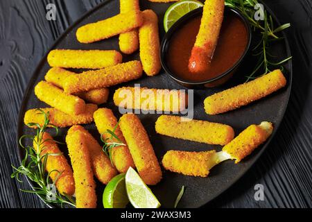 gros plan de bâtonnets de mozzarella frits avec sauce, tranches de citron vert et romarin frais sur un plateau noir sur une table en bois noir Banque D'Images