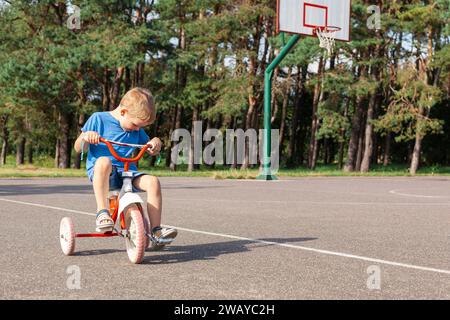 Un petit garçon monte un tricycle dans un terrain de basket-ball de ville, il est pédales de pédale et regarde vers le bas. Banque D'Images