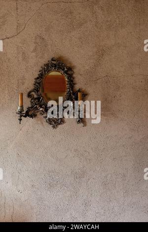 Un petit miroir ovale antique avec un cadre en cuivre et des chandeliers sur un vieux mur de plâtre brun fissuré. Banque D'Images